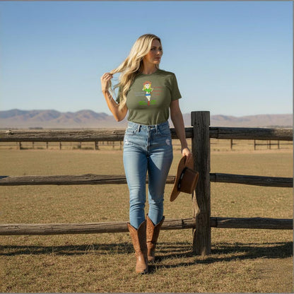 Woman wearing a green t-shirt with colorful cowgirl graphics and text, standing by a wooden fence in a rural landscape wearing a green t-shirt and blue jeans.