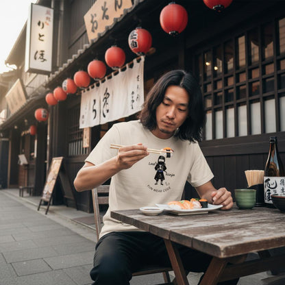 Man wearing a beige T-shirt with a playful "Last Ninja" graphic design and text "FUN WEAR CODE", eating sushi outdoors in a traditional Japanese setting with lanterns and wooden architecture.