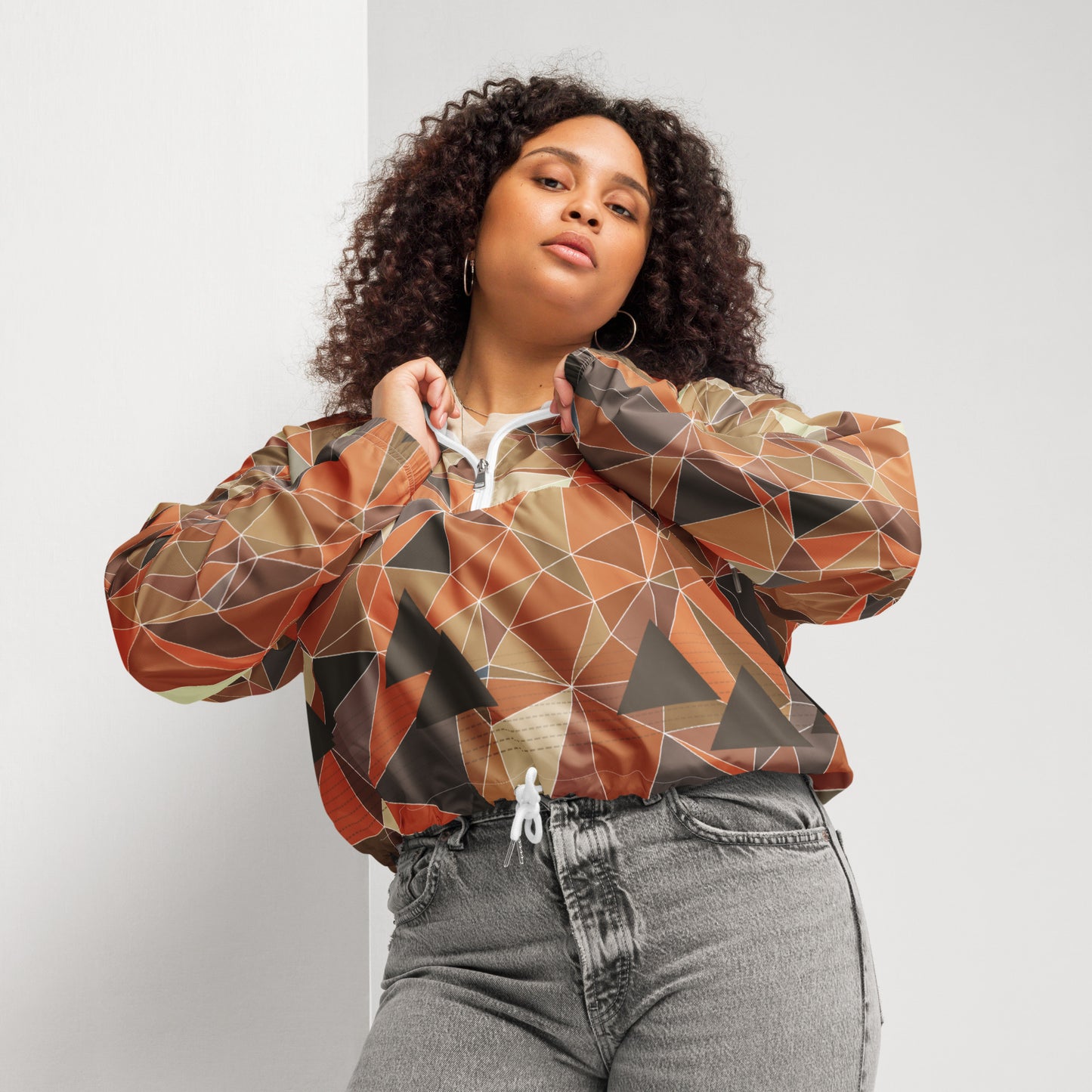 Woman wearing a cropped multicolor geometric patterned windbreaker jacket and gray jeans against a plain background