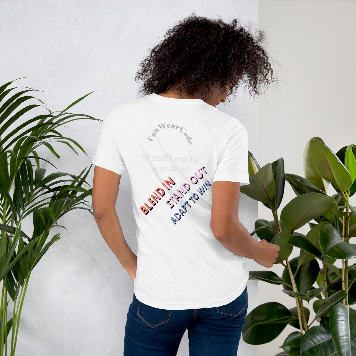 Person wearing a white t-shirt with a 'Fun Wear Code' text logo and colorful texts on the back, standing between two plants against a white wall.